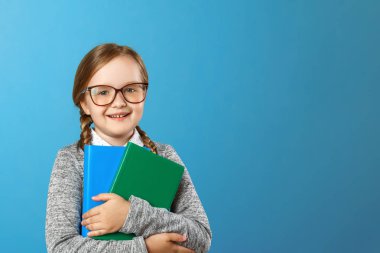 Portrait of a charming little girl schoolgirl in glasses on a blue background. The child holds books. The concept of education and school. Copy space