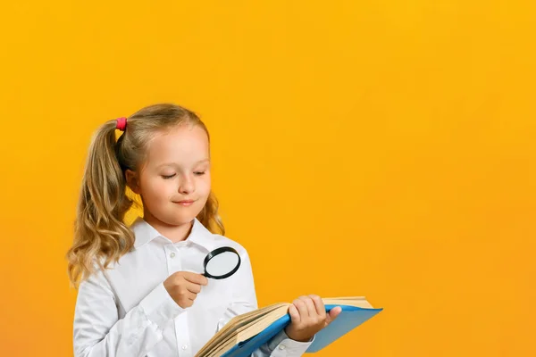 La niña está leyendo un libro sobre la mesa con una lupa sobre un fondo amarillo. El concepto de educación. Regreso a la escuela — Foto de Stock