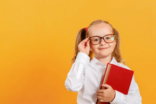 Ritratto di una bambina carina su sfondo giallo. Studentessa bambino guardando la fotocamera, tenendo un libro e raddrizza i bicchieri. Il concetto di istruzione. Copia spazio — Foto Stock