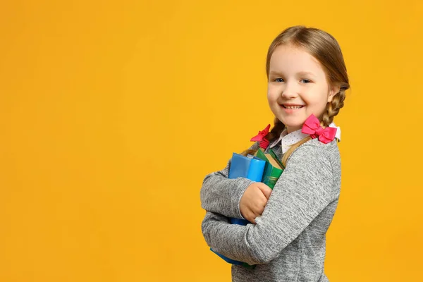 Ritratto di una scolaretta con le trecce su sfondo giallo. Il bambino sorride e tiene libri di testo. Il concetto di istruzione. Torniamo a scuola. Copia spazio — Foto Stock