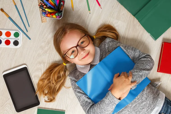 Yerde yatan bir kitapla gözlüklü sevimli küçük bir kız. Bir çocuk bir kitap, tablet, boyalar, fırçalar, kalemler ile çevrilidir.