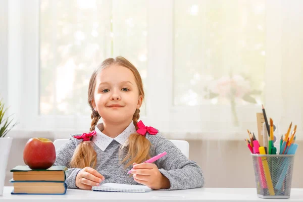 Una studentessa seduta al tavolo. Studentessa con una penna in mano e con un quaderno, il bambino guarda nella fotocamera. Il concetto di istruzione e scuola. Sfondo sfocato . — Foto Stock