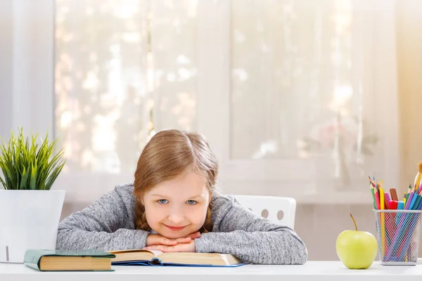 Allegro bambina studentessa è seduto al tavolo, mettere la testa nelle sue mani e guarda nella fotocamera. Il concetto di istruzione e scuola. Sfondo sfocato . — Foto Stock