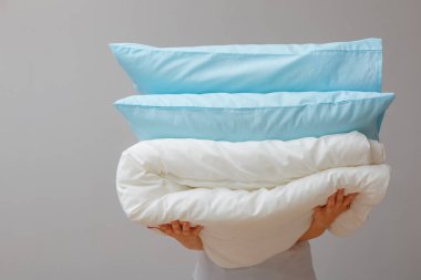 Gri duvarlı bir arka plan üzerinde yorgan ve yastık tutan kadın. Uyumak için yatak yığını. Ev.