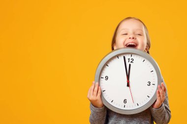 Küçük bir kız büyük saatin altından dışarı bakıyor. Sarı arka plan. Eğitim kavramı, okul, öğrenme zamanı.