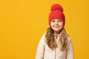 Bej ceketli ve sarı arka plan üzerinde kırmızı bir şapka giyen neşeli bir çocuğun portresi. Küçük kız sarışın kameraya bakar. Sonbahar konsepti. Kopyalama alanı