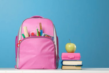 Öğrenci seti. Kırtasiyeli pembe sırt çantası, bir yığın kitap, bir beslenme çantası, mavi arka plan üzerinde masanın üzerinde bir elma. Okula geri dön.