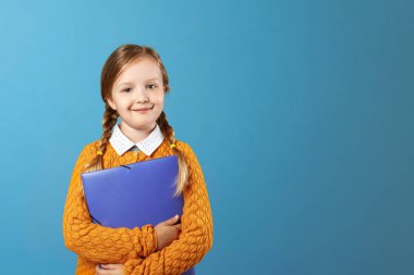 Küçük bir kız öğrencinin yakın çekim portresi. Sarı kazak güzel çocuk mavi bir arka plan üzerinde kağıtları için mor bir klasör tutar. Kopyalama alanı