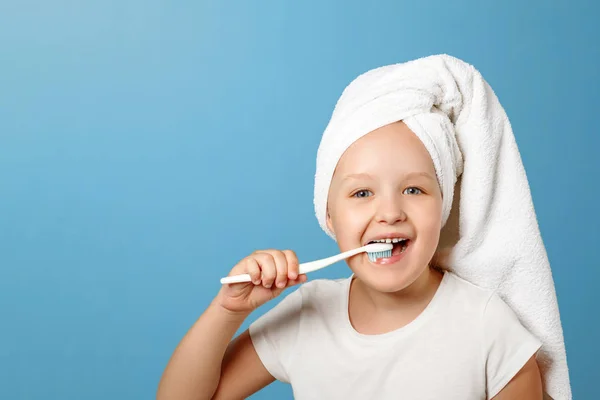 Je to portrét malé holčičky na modrém pozadí. Dítě s bílým ručníkem na hlavě si otíká zuby. Pojetí každodenní hygieny — Stock fotografie