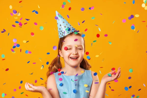Cheerful little girl celebrates birthday. The child is standing with eyes closed in the rain of confetti. Closeup portrait on yellow background. — Stock Photo, Image