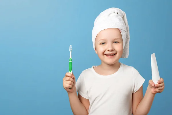 Je to portrét malé holčičky na modrém pozadí. Dítě s bílým ručníkem na hlavě, držící zubní kartáček a zubní pastu. Pojem každodenní hygieny. Kopírovat místo — Stock fotografie
