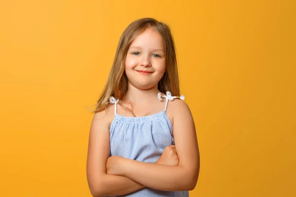 Portrét směšné malé blonďaté dívky na žlutém pozadí. Dítě složila paže a dívá se do kamery. — Stock fotografie
