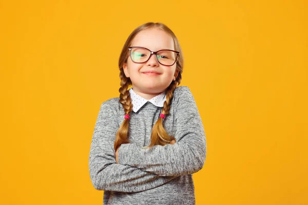 Portrét veselé holčičky v brýlích na žlutém pozadí. Dětská školka složila ruce a dívá se do kamery. — Stock fotografie