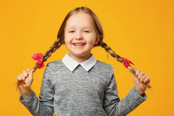 Sarı arka planda neşeli bir küçük kızın yakın çekim portresi. Çocuk kız öğrenci güler, ellerini pigtails tutar ve kameraya bakar.