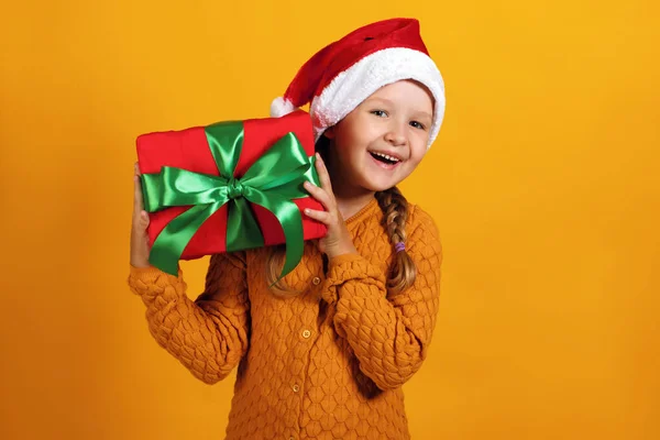 Felice bambina in cappello di Babbo Natale tiene regalo di Natale su sfondo giallo — Foto Stock