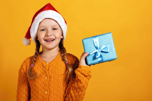 Felice bambina in cappello di Babbo Natale tiene scatola di Natale con regalo su sfondo giallo — Foto Stock
