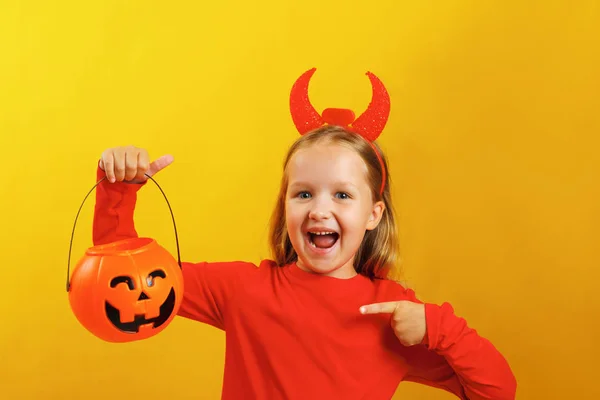 Halloween celebration. Little girl in a devil costume on a yellow background. The child holds a pumpkin jack lantern and points a finger at the bucket — Stock Photo, Image