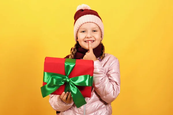 Ritratto di una graziosa bambina in giacca, sciarpa e cappello su sfondo giallo. Il bambino tiene una scatola con un regalo e mostra un segno shh — Foto Stock