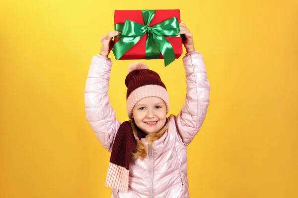 Ritratto di una graziosa bambina in giacca, sciarpa e cappello su sfondo giallo. Il bambino tiene una scatola con un regalo sopra la testa . — Foto Stock