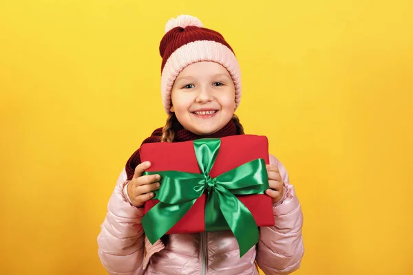 Niña feliz en un sombrero y bufanda con una caja de regalo sobre un fondo amarillo . — Foto de Stock