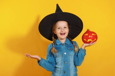 Sarı bir arka plan üzerinde bir cadı şapka neşeli küçük bir kızın closeup portre. Çocuk balkabağı jack fener tutar. Cadılar Bayramı kutlaması