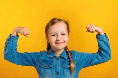 Sarı bir arka plan üzerinde saç yapılmış pigtails ile kot gömlek li küçük kız. Çocuk güçlü eller gösterir. Eğitim kavramı, okul, başarı, güç, feminizm
