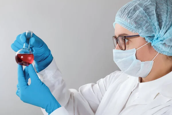 Num laboratório de química, uma farmacêutica está a analisar um frasco com líquido vermelho. Experiência médica com um tubo. Químico trabalhando em um teste com luvas azuis, roupas de proteção e óculos — Fotografia de Stock