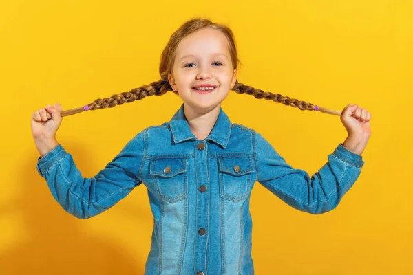 Portrét veselé holčičky v džínovou košili na žlutém pozadí. Dítě má ruce s copánky a dívá se do kamery — Stock fotografie