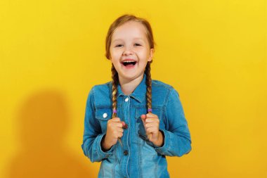 Sarı bir arka plan üzerinde bir kot gömlek neşeli mutlu küçük kız. Çocuk güler ve saç örgüleri tutar