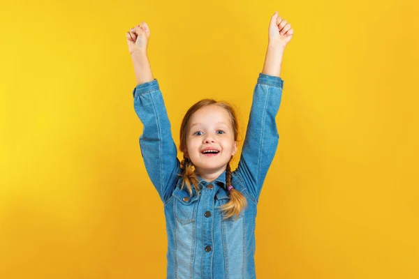 Cheerful happy little girl raised her hands up. A child in a denim shirt on a yellow background — Stock Photo, Image