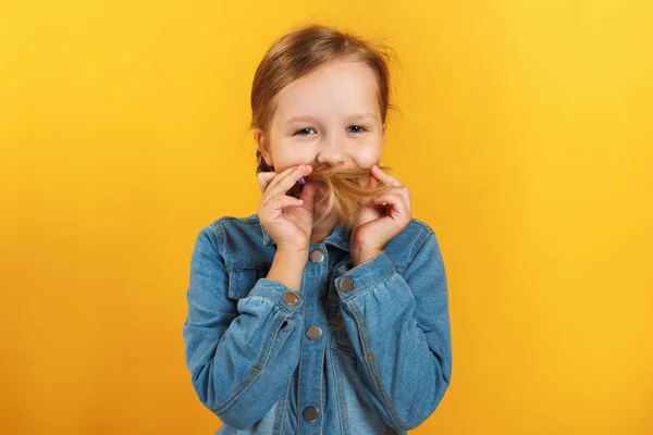 Šťastná holčička si z vlasů dělá knírek. Dítě s džínovou košilí na žlutém pozadí — Stock fotografie