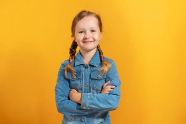 Sarı arka plan üzerinde kot gömlekli küçük bir kız. Çocuk kollarını çapraz ve gülümsüyor.