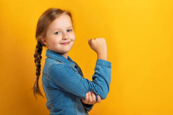 Je to portrét malé holčičky na žlutém pozadí. Dětská ruka ukazuje biceps. Koncepce síly dívek. — Stock fotografie