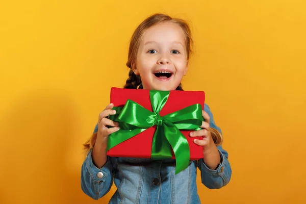 Retrato de una linda niña feliz en una camisa de mezclilla sobre un fondo amarillo. El niño sostiene una caja con un regalo en la mano — Foto de Stock