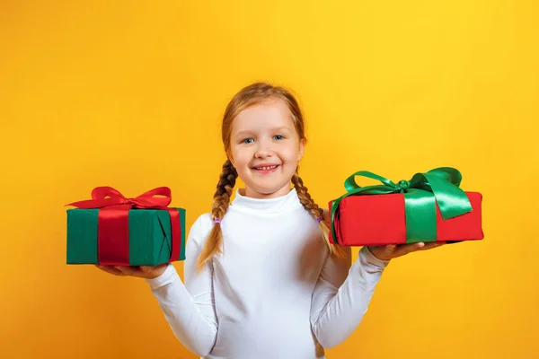 Retrato de una niña feliz con un cuello alto blanco sobre un fondo amarillo. El niño sostiene cajas con regalos en sus manos. — Foto de Stock