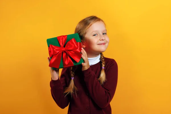 Ritratto di una graziosa bambina in maglione rosso su sfondo giallo. Il bambino tiene una scatola con un regalo — Foto Stock