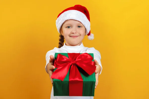 Ragazzina felice con un regalo per Natale. Un bambino con un cappello di Natale fa un regalo su uno sfondo giallo — Foto Stock