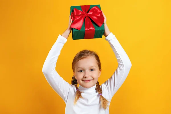 Linda niña feliz sosteniendo una caja con un regalo sobre su cabeza. Un niño con coletas en cuello alto blanco sobre un fondo amarillo — Foto de Stock