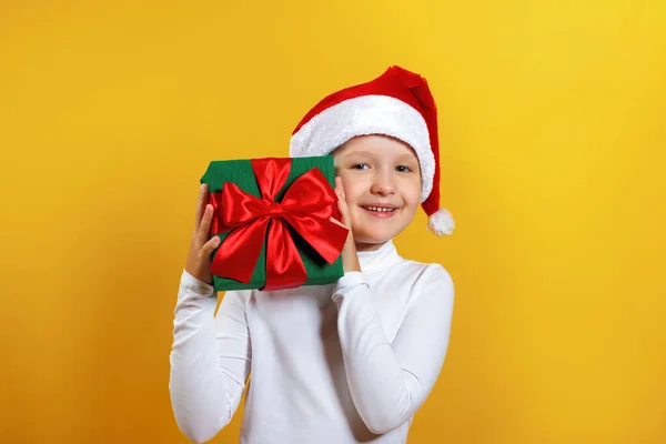 Bonne petite fille dans le chapeau de Père Noël détient cadeau de Noël. Un enfant au col roulé blanc sur fond jaune — Photo