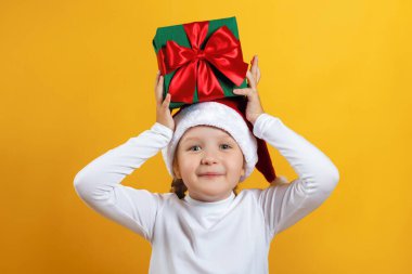 Sarı arka planda noel hediyesi olan mutlu küçük kız. Şapkalı bir çocuk kafasında bir hediye taşıyor.