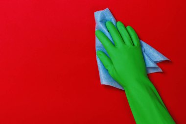 Kırmızı arka planda mavi bir bez parçası olan koruyucu yeşil eldivenli bir işçi eli. Temizlik kavramı