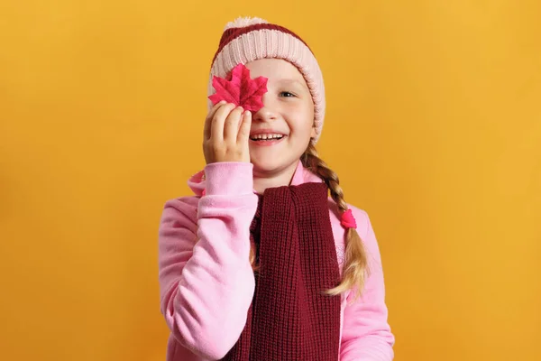 Šťastná holčička drží javorový list. Dítě v šátku a klobouku na žlutém pozadí. Podzimní koncept — Stock fotografie