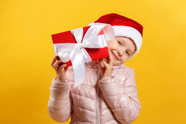 Ragazzina felice in cappello di Babbo Natale sta tenendo regalo di Natale. Bambino in giacca rosa su sfondo giallo — Foto Stock