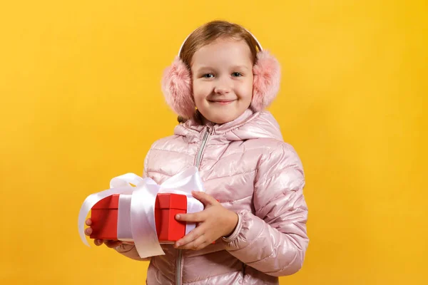 Ragazzina felice tiene una scatola con un regalo. Un bambino in giacca e polsini per le orecchie caldi su uno sfondo giallo. Natale e nuovo concetto di anno — Foto Stock