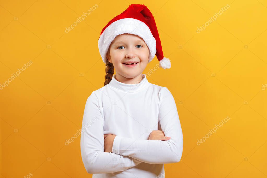 Charming portrait of a happy cute little girl in santa hat. The child crossed his arms and looks at the camera on a yellow background