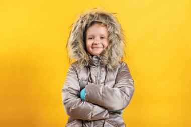 Sarı arka planda ceketli mutlu küçük kız. Çocuk kollarını kavuşturdu ve kameraya baktı. Kış konsepti