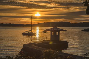 Bir körfezin üzerinde gün batımında yelkenli bir tekne ve iskelede küçük bir ev..