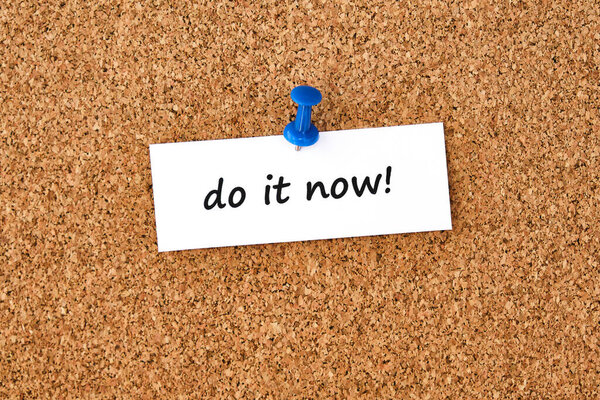 Do it now. Text written on a piece of paper or note, cork board background.