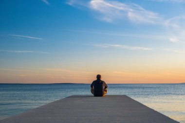 Kapüşonlu ve kısa kargolu bir adam rıhtımın kenarında oturmuş meditasyon yapıyor. Deniz ufku, Pag adası, Adriyatik denizi, Hırvatistan