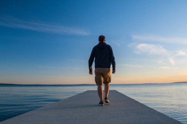 Kapüşonlu adam ve limanda kısa kargo pantolonu, arka planda deniz ufku, Pag Adası, Adriyatik denizi, Hırvatistan